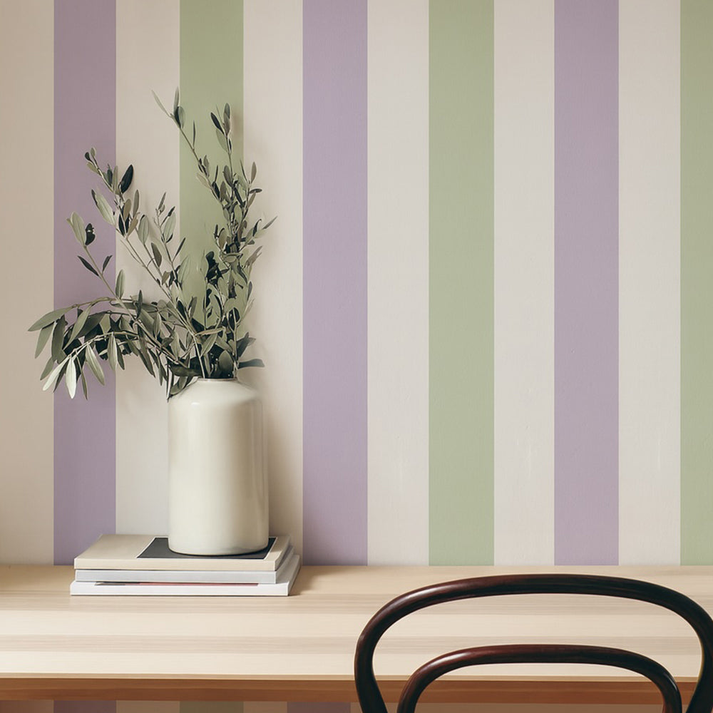 A sleek, off-white ceramic vase with a smooth, cylindrical body holds delicate olive branches against a backdrop of soft lavender and sage green vertical stripes.