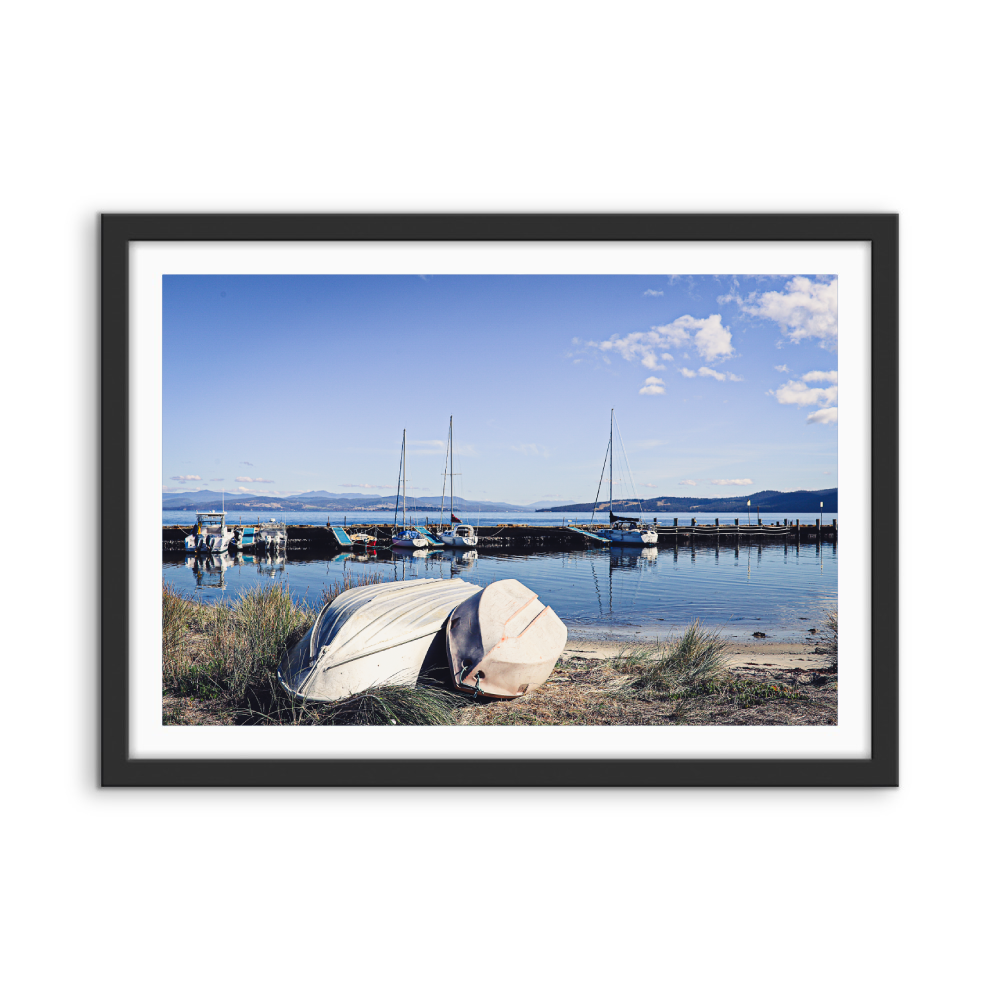 Black-framed art print of a serene harbor scene with boats and calm water under a blue sky by edwina cameron