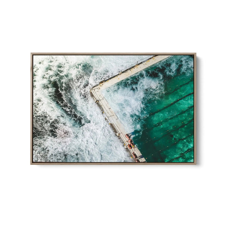 A framed fine art print of a vibrant turquoise ocean pool with white waves and a concrete walkway in a sleek metallic frame