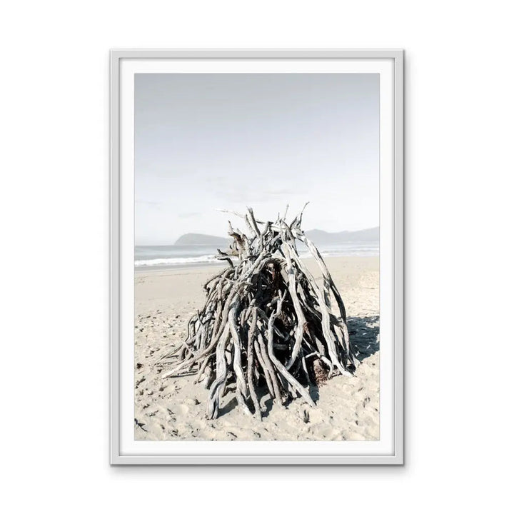 A framed photographic print of weathered driftwood on a sandy beach under a soft sky by Edwina Cameron