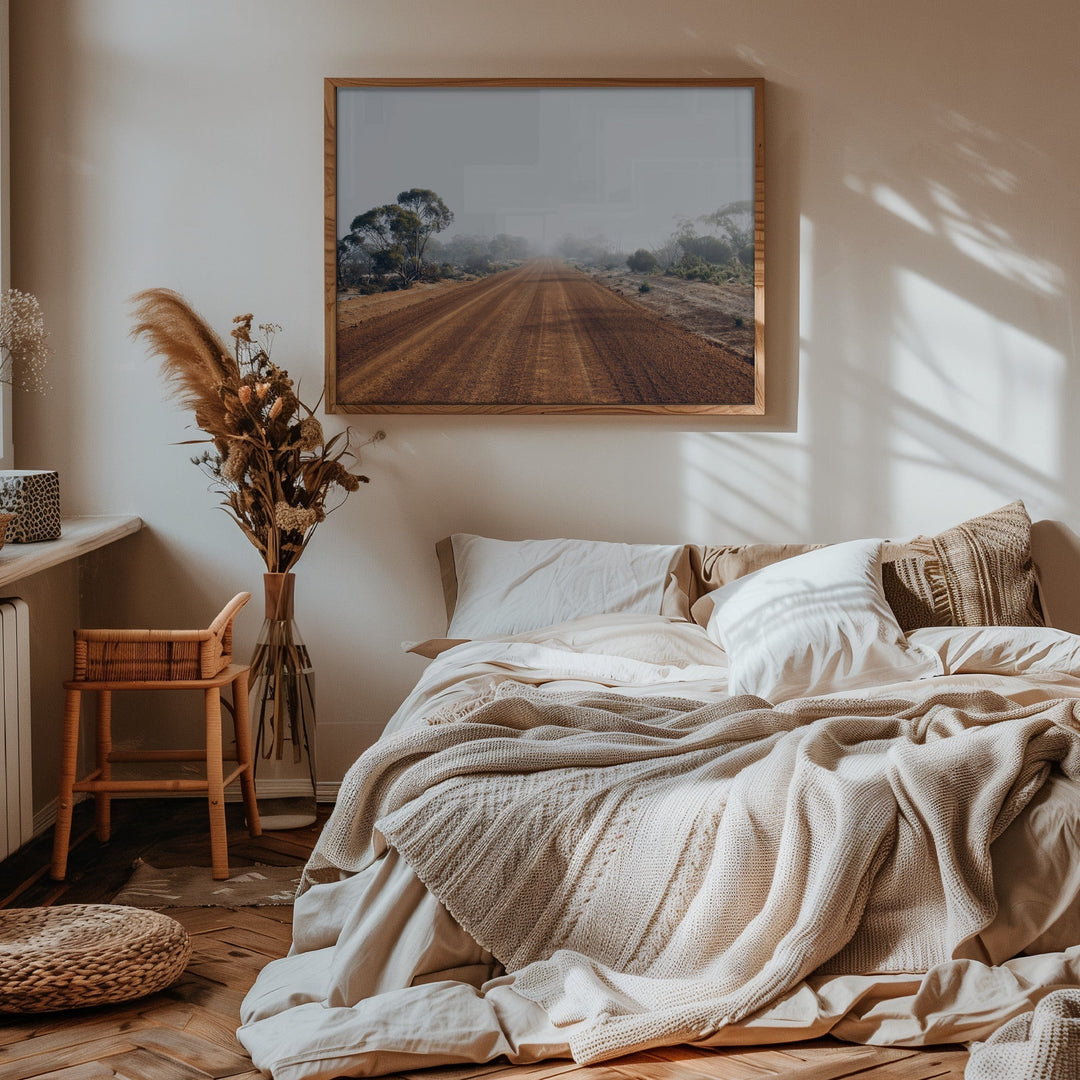 A rustic wooden-framed photographic print of a misty dirt road landscape from the australian outback mounted above a bed