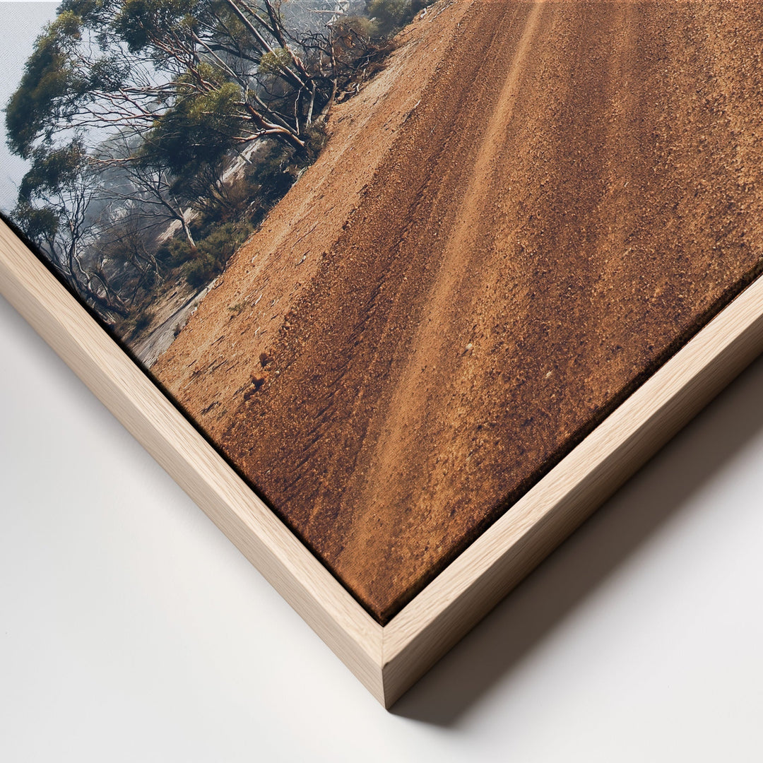 A rustic wooden-framed canvas print of a red dirt road winding through trees in earthy brown tones australian outback landscape