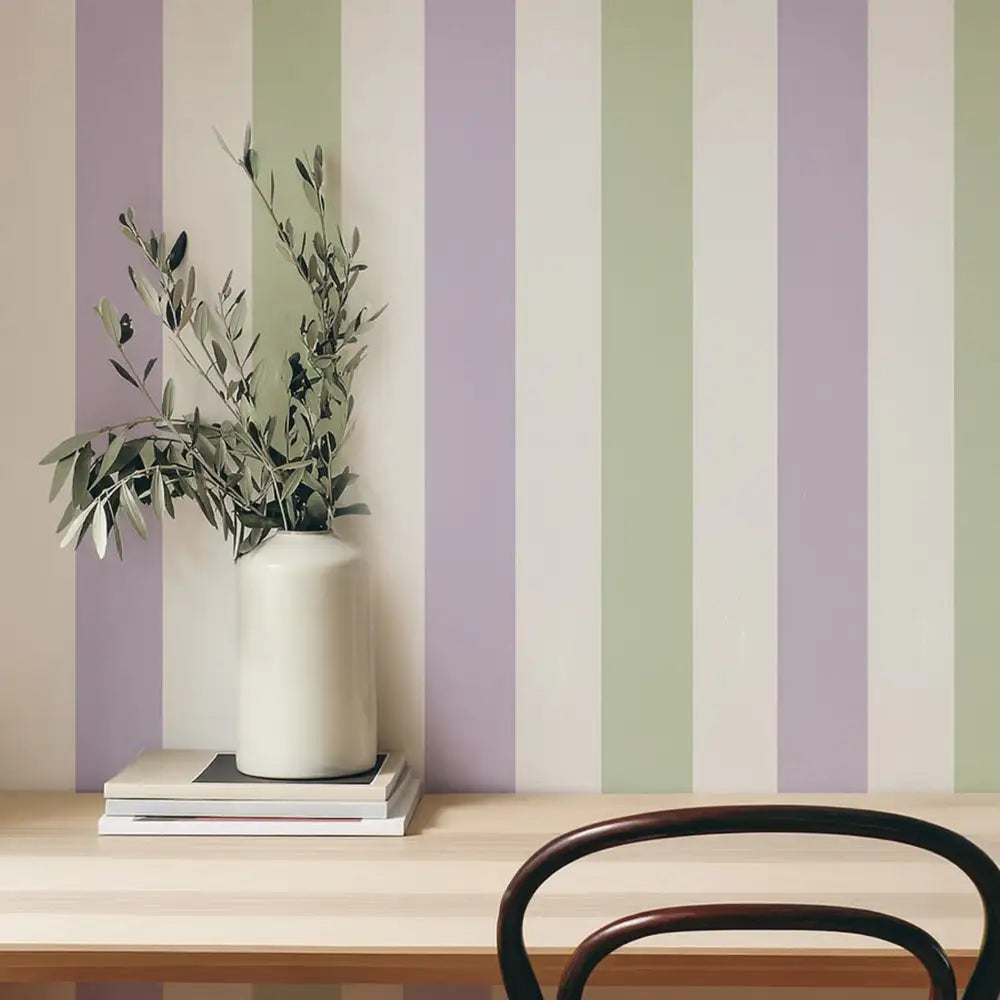 A sleek, off-white ceramic vase with a smooth, cylindrical body holds delicate olive branches against a backdrop of soft lavender and sage green vertical stripes.