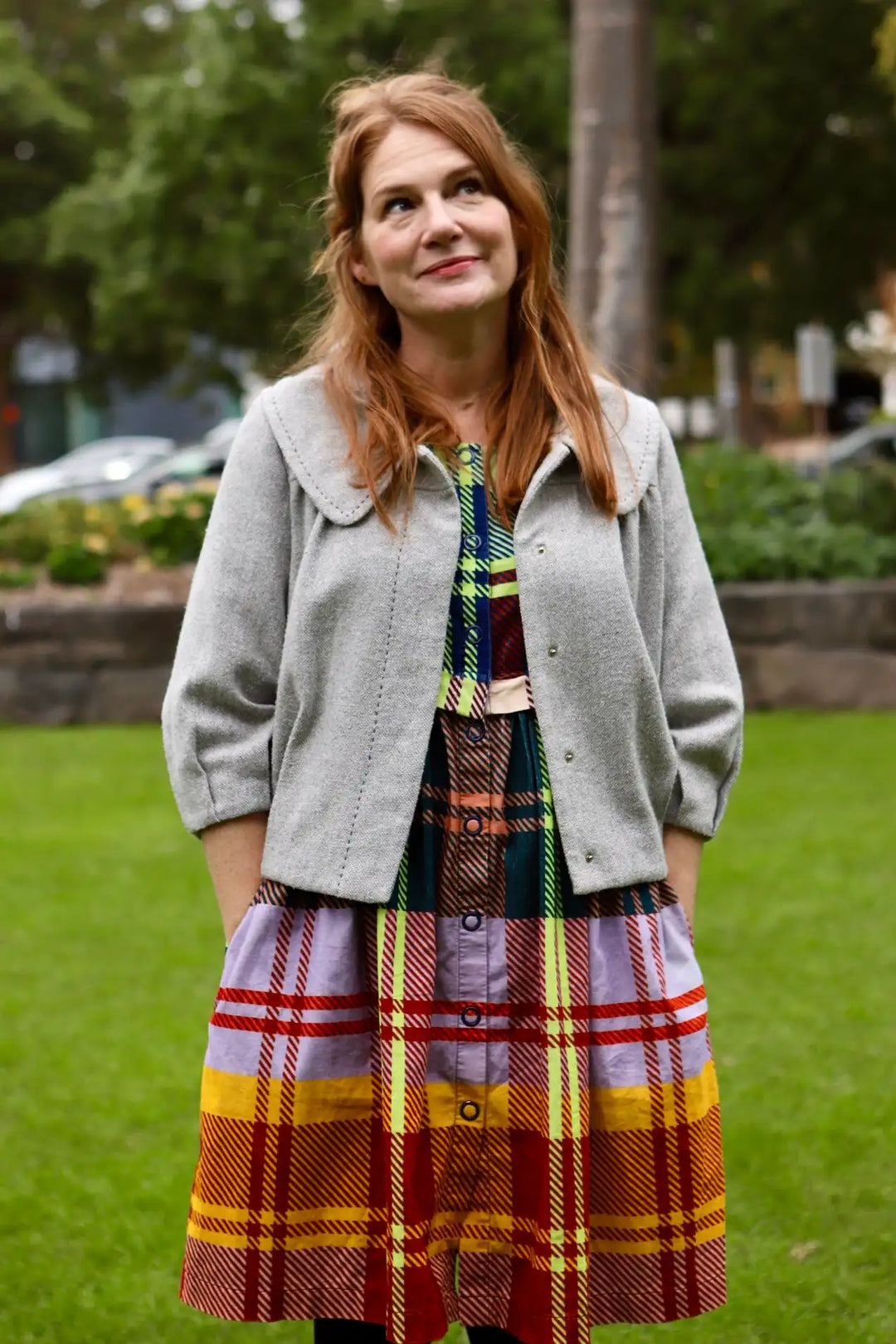 A woman wears a gray woolen cardigan over a multi-colored plaid dress with bold red, yellow, and purple stripes.